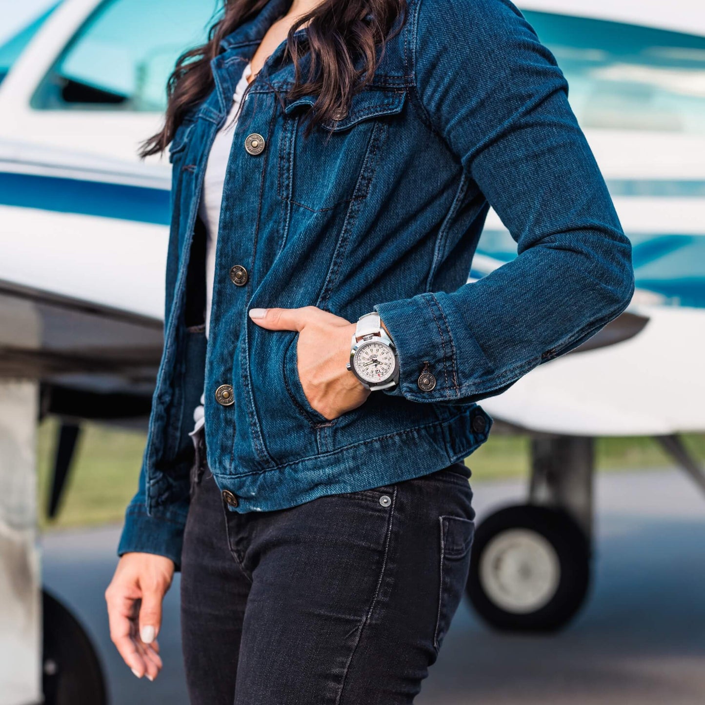 Amelia Women's Pilot Watch with Cloud White Dial and White Leather Strap, Worn by Woman in Denim Jacket Standing Beside Airplane on Tarmac