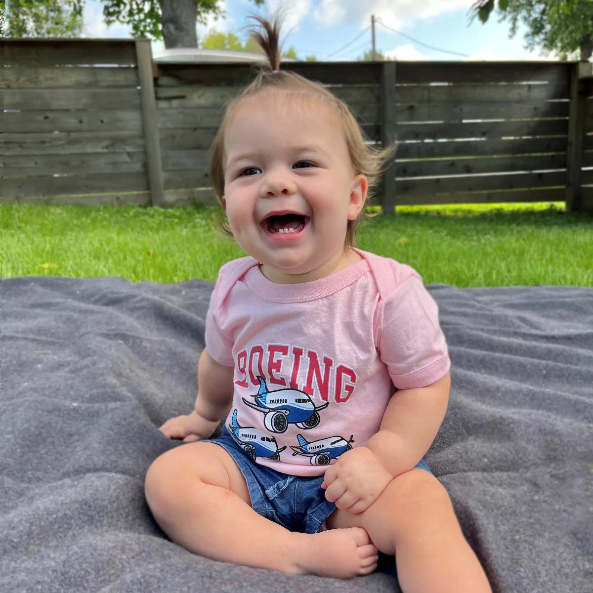 Baby wearing pink Boeing Baby Varsity Pudgy Onesie with airplane graphics while sitting outdoors