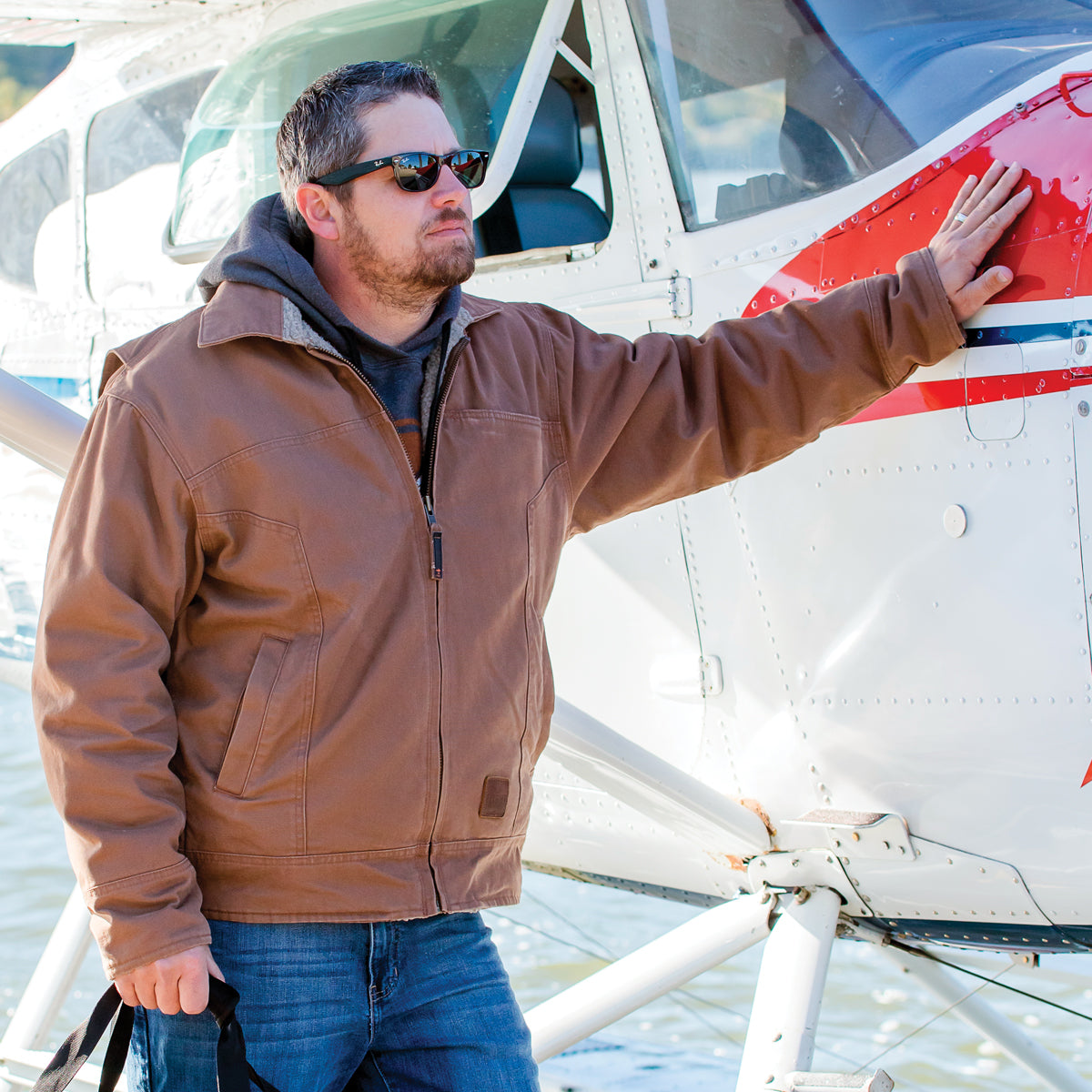 Man wearing sunglasses and Brown Flight Outfitters Bush Pilot Jacket with hand on seaplane cowl