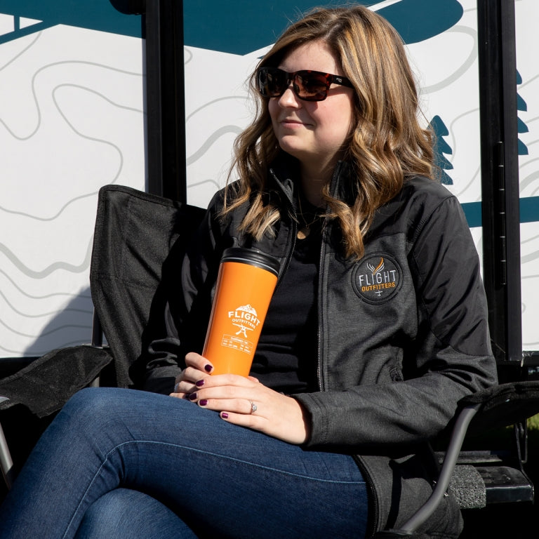 Lifestyle photo – woman sitting outdoors by RV wearing Flight Outfitters Women's Kodiak Jacket with logo, holding orange tumbler